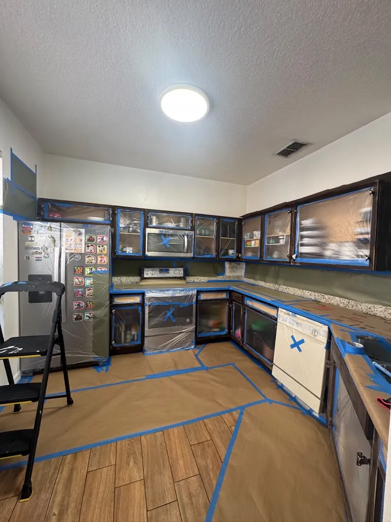 A kitchen with dark cabinets, doors off, floor papered and every opening wrapped in plastic before painting