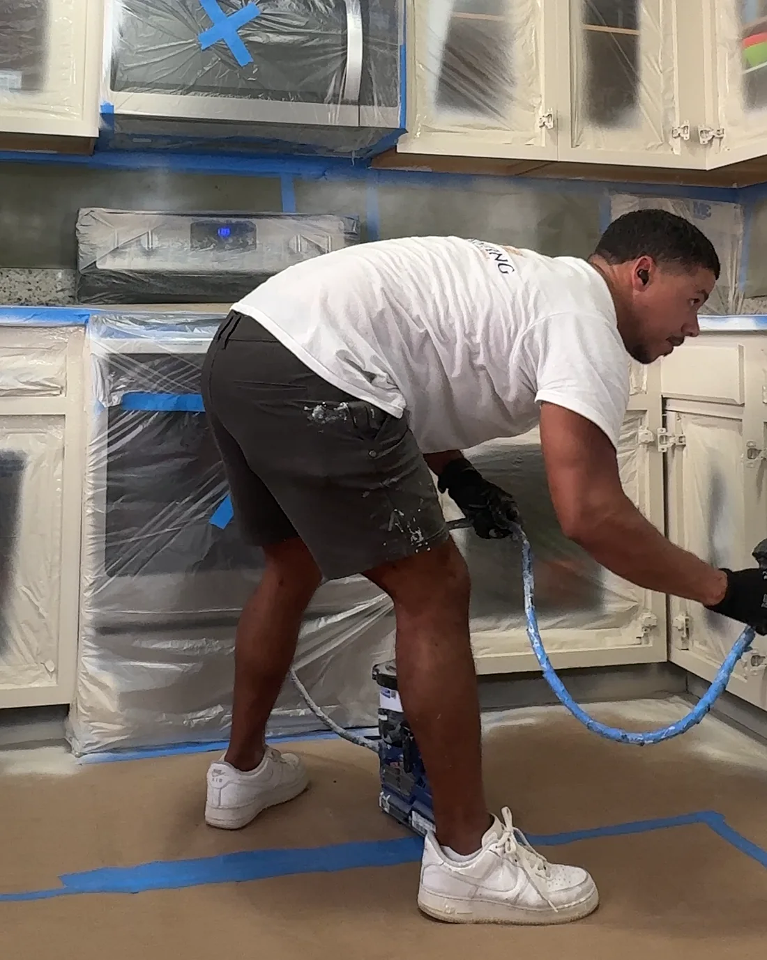 Leandro spraying the lower cabinets in a kitchen covered in plastic and paper